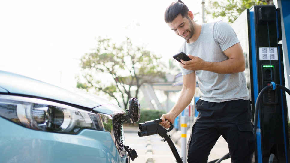 Egy férfi elektromos autóját tölti egy nyilvános töltőállomáson, miközben a telefonját használja.